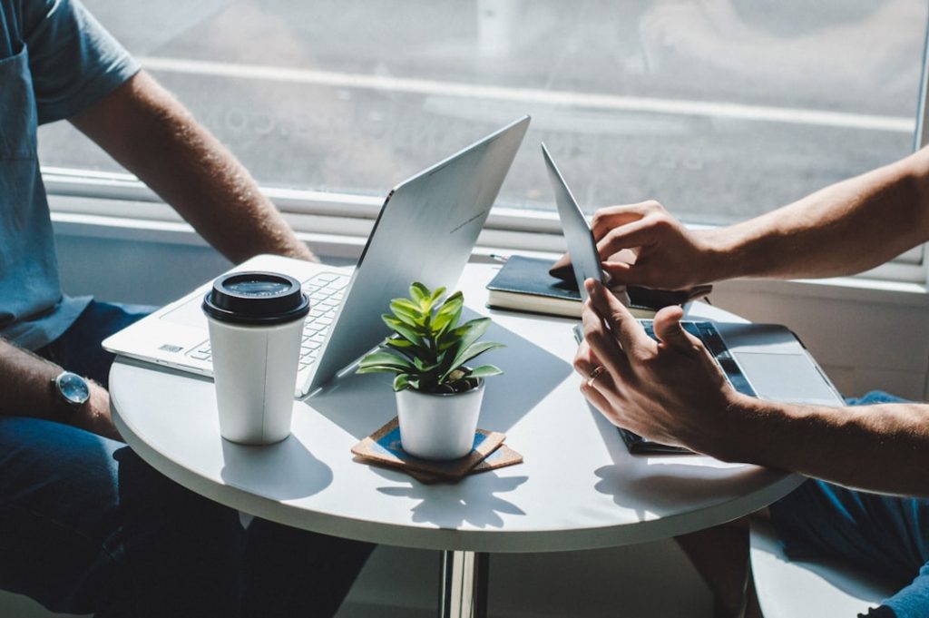 Two people working side by side at a small round cafe table in bright window light, with open laptops, a takeaway coffee cup, a notebook and a potted plant between them.