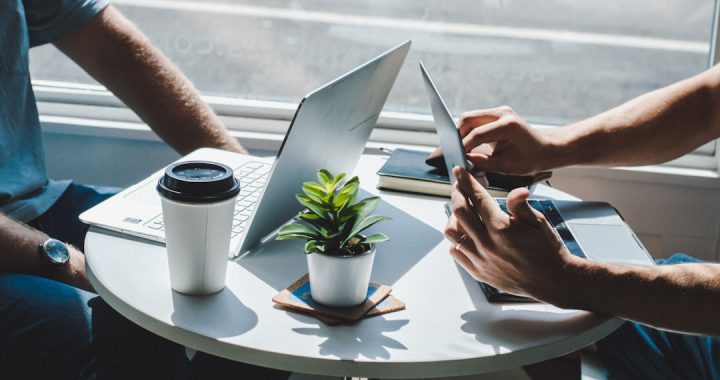 Two people working side by side at a small round cafe table in bright window light, with open laptops, a takeaway coffee cup, a notebook and a potted plant between them.