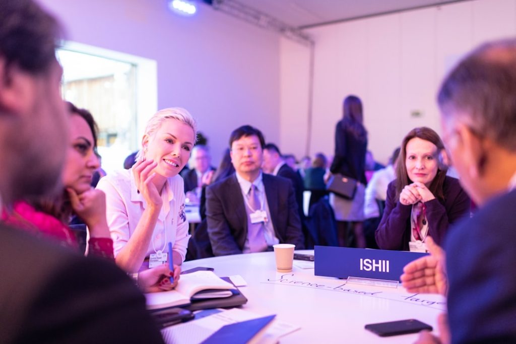 Professionals seated around a white table at a networking event, wearing name badges and talking mid-conversation, with notebooks and a table nameplate in front of them.