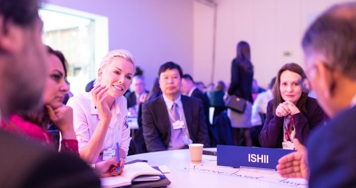 Professionals seated around a white table at a networking event, wearing name badges and talking mid-conversation, with notebooks and a table nameplate in front of them.