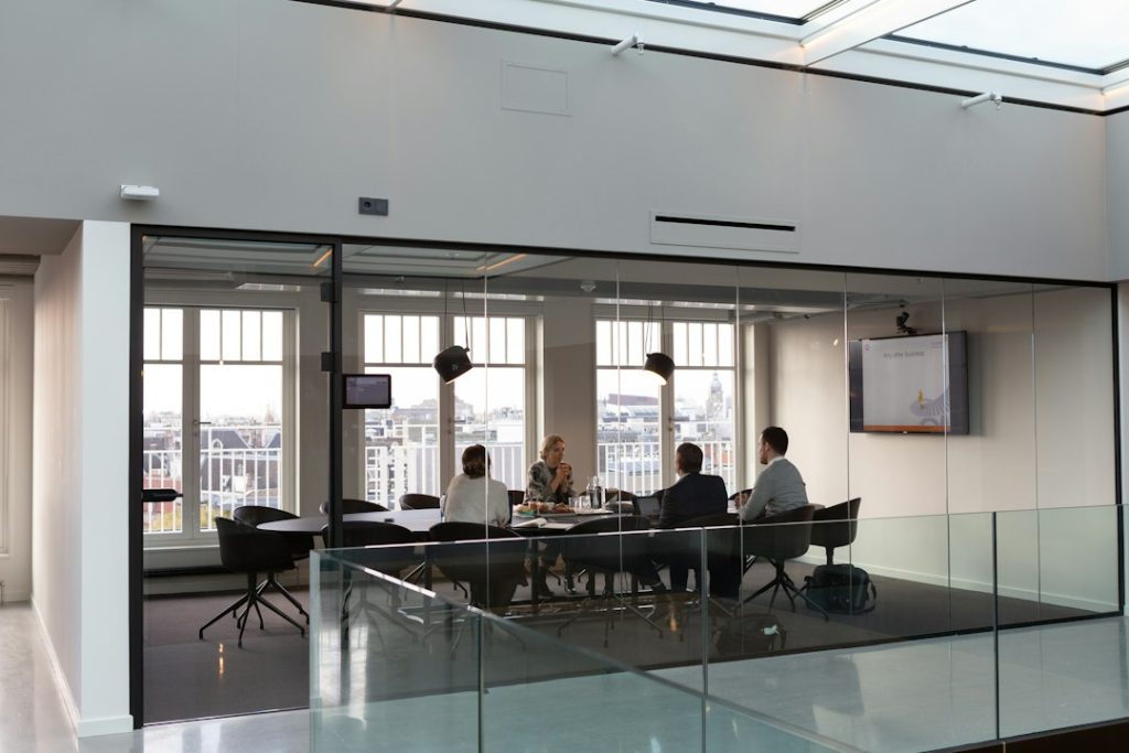 Four people seated around a black table in a glass-walled conference room, seen from outside the glass, with tall windows behind them looking out over a city rooftop skyline and a wall-mounted screen showing a presentation slide.