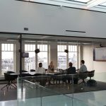 Four people seated around a black table in a glass-walled conference room, seen from outside the glass, with tall windows behind them looking out over a city rooftop skyline and a wall-mounted screen showing a presentation slide.