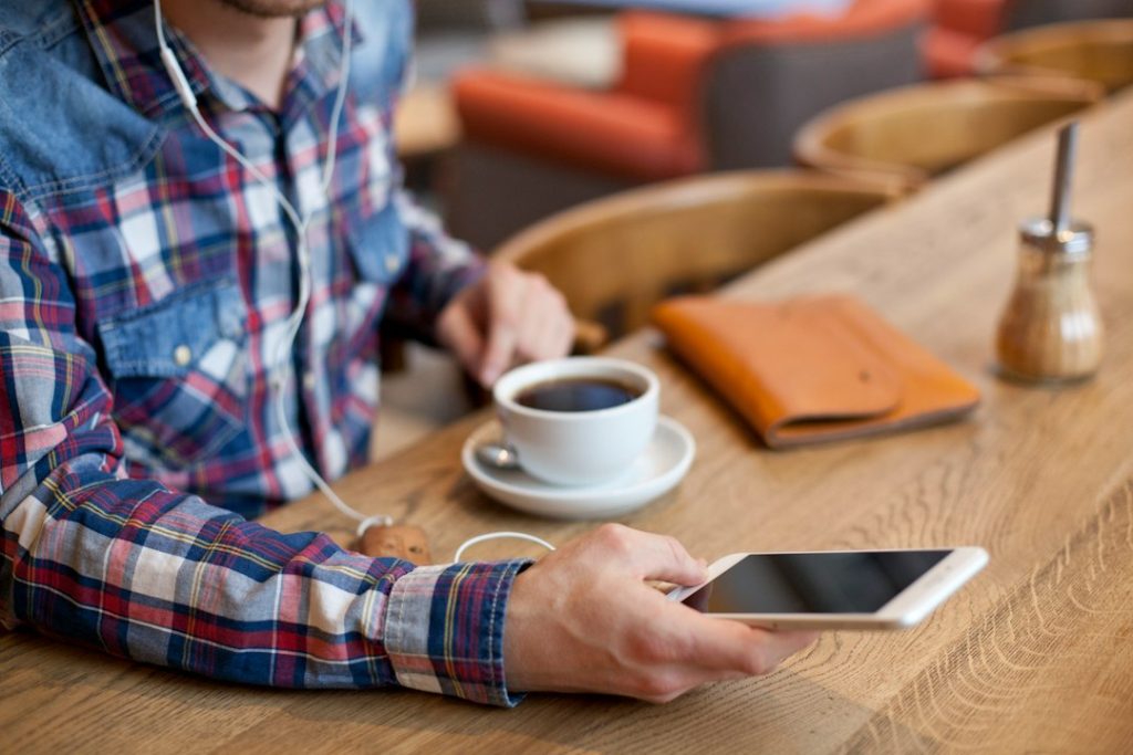 A man in a plaid shirt with earbuds sits at a wooden café counter holding a smartphone, a cup of black coffee and a tan leather notebook beside him.