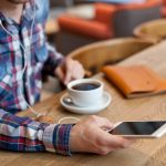 A man in a plaid shirt with earbuds sits at a wooden café counter holding a smartphone, a cup of black coffee and a tan leather notebook beside him.