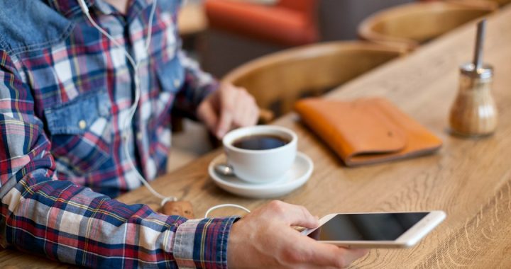 A man in a plaid shirt with earbuds sits at a wooden café counter holding a smartphone, a cup of black coffee and a tan leather notebook beside him.