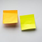 Two blank sticky notes, one orange and one yellow, stuck side by side on a plain white wall.