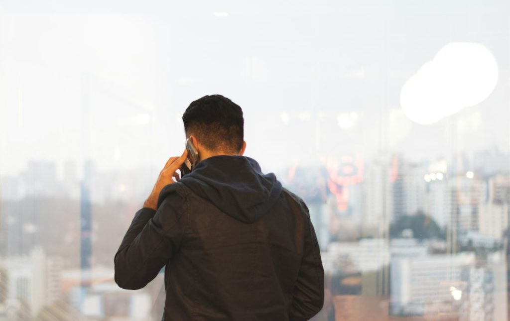 Rear view of a dark-haired man in a black jacket with a navy hood, holding a phone to his ear while standing at a floor-to-ceiling office window overlooking a hazy city skyline of high-rise buildings.