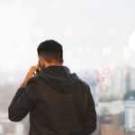 Rear view of a dark-haired man in a black jacket with a navy hood, holding a phone to his ear while standing at a floor-to-ceiling office window overlooking a hazy city skyline of high-rise buildings.