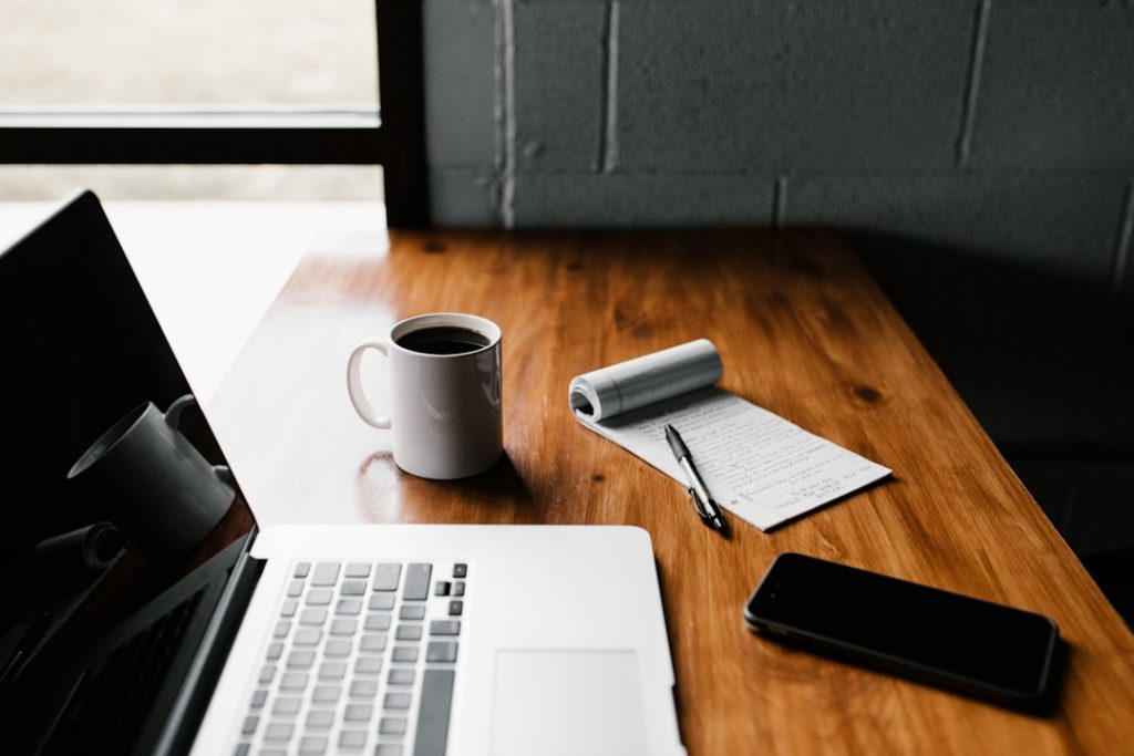 An open laptop with a dark screen sits on a wooden desk by a window, next to a white mug of black coffee, a flipped-open notepad covered in handwritten notes with a pen laid across it, and a phone face down on the desk.