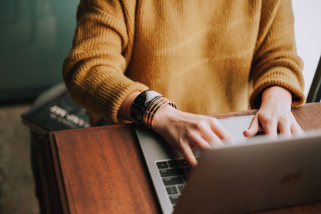A person in a mustard-yellow knit sweater typing on a silver laptop at a wooden desk, with a stack of books beside them and no screen content visible.