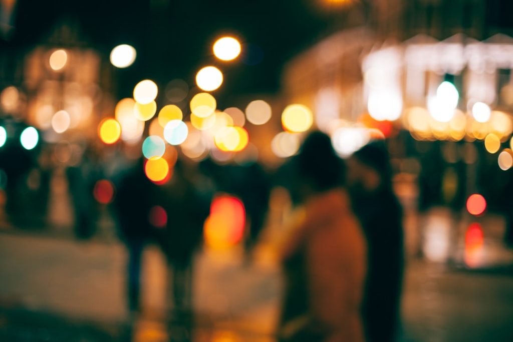 A city street at night photographed completely out of focus, so the dozen people crossing it read only as dark silhouettes against orange, red and white circles of blurred streetlight and shopfront glow. Movement is obvious; not one face is identifiable.