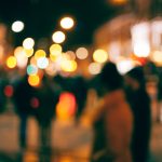 A city street at night photographed completely out of focus, so the dozen people crossing it read only as dark silhouettes against orange, red and white circles of blurred streetlight and shopfront glow. Movement is obvious; not one face is identifiable.
