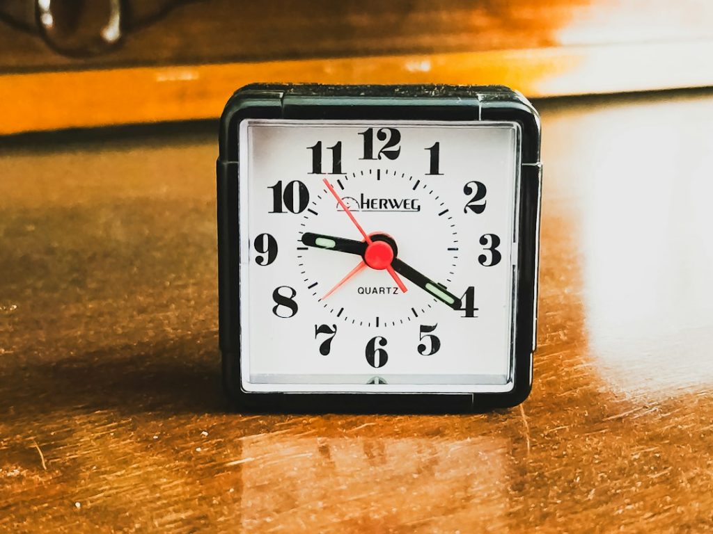 A small square black alarm clock standing on a scratched wooden surface, its white face showing the time at roughly twenty past eight, with a red second hand and warm orange light reflecting off the wood behind it.