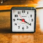 A small square black alarm clock standing on a scratched wooden surface, its white face showing the time at roughly twenty past eight, with a red second hand and warm orange light reflecting off the wood behind it.