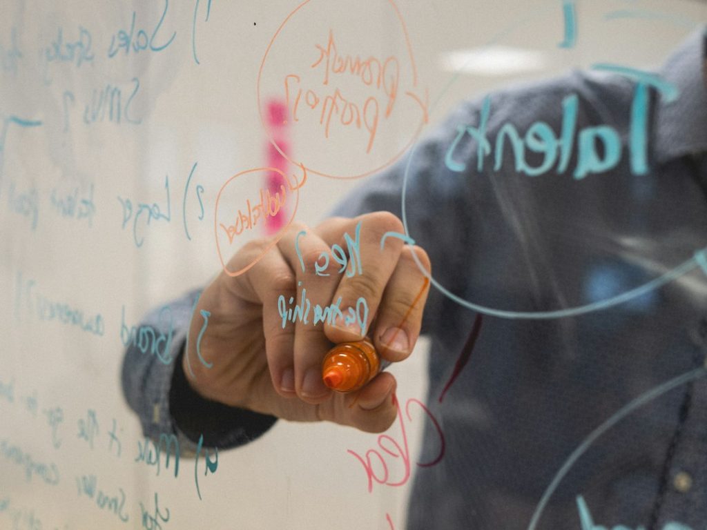 A person in a denim shirt writes with an orange marker on a glass wall, circling and listing planning notes in blue and orange ink, seen in reverse through the glass.