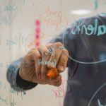 A person in a denim shirt writes with an orange marker on a glass wall, circling and listing planning notes in blue and orange ink, seen in reverse through the glass.