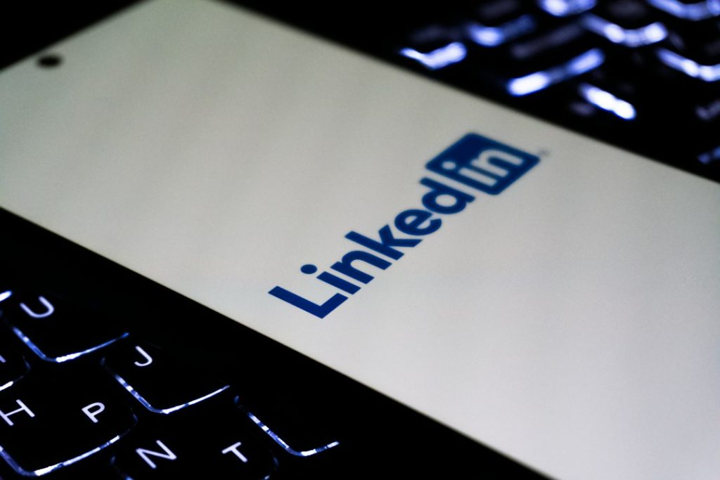 Smartphone lying on a backlit laptop keyboard, its screen showing the blue LinkedIn logo on a white background
