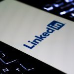 Smartphone lying on a backlit laptop keyboard, its screen showing the blue LinkedIn logo on a white background