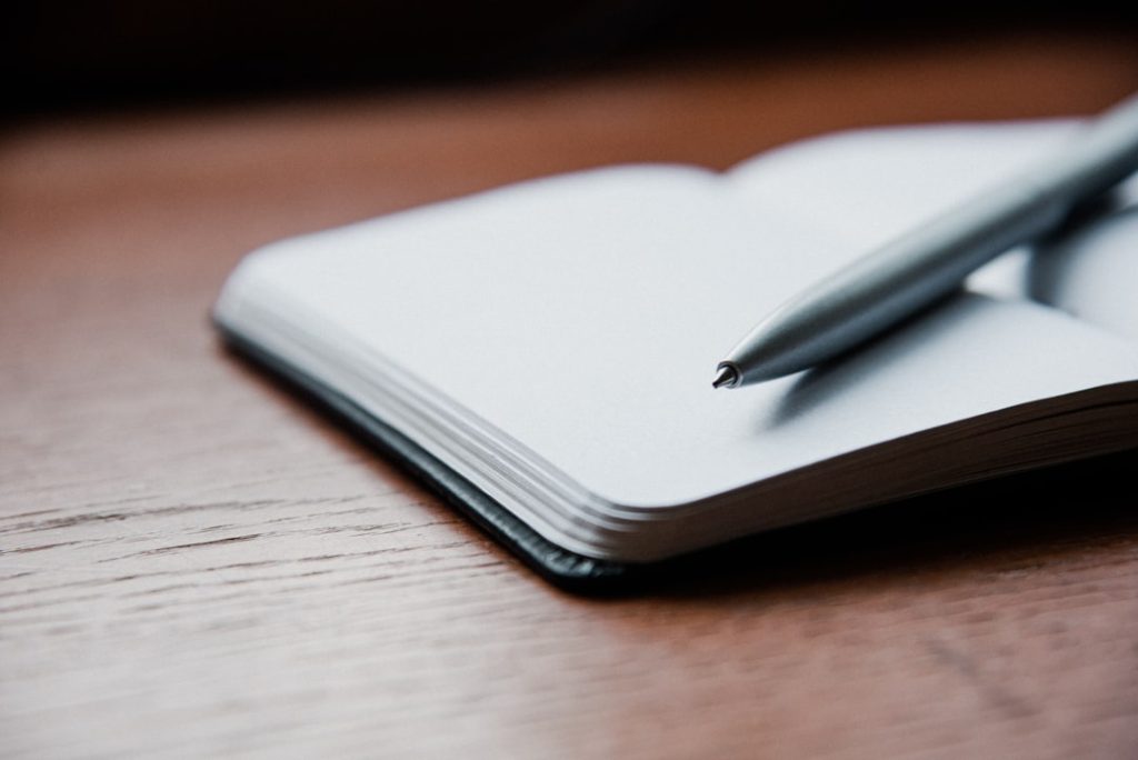 An open notebook with blank pages and a silver ballpoint pen resting across it on a wooden desk.