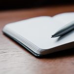 An open notebook with blank pages and a silver ballpoint pen resting across it on a wooden desk.