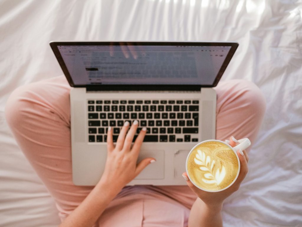 Overhead view of a person sitting cross-legged on a white bed, typing a document on a laptop while holding a latte with leaf-pattern foam art.