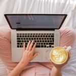 Overhead view of a person sitting cross-legged on a white bed, typing a document on a laptop while holding a latte with leaf-pattern foam art.