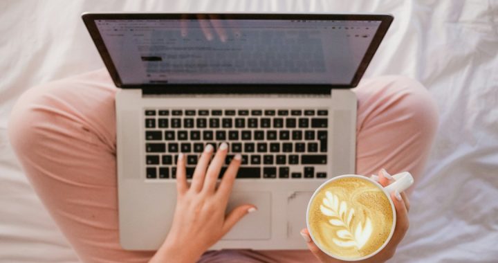Overhead view of a person sitting cross-legged on a white bed, typing a document on a laptop while holding a latte with leaf-pattern foam art.