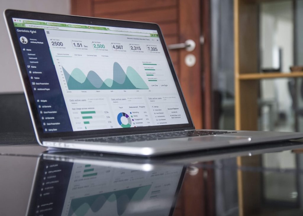 An open silver laptop on a reflective glass table, its screen filled with a web analytics dashboard: a row of counter tiles reading 2,500 total users, 1.51 second average time, 2,500 total mails, 4,567 templates, 2,315 collections and 7,325 total connections, above a teal area chart running January through June and, below it, green bar charts and a multicolour donut chart breaking activity into segments. A wooden door and a glass office partition sit out of focus behind the screen.