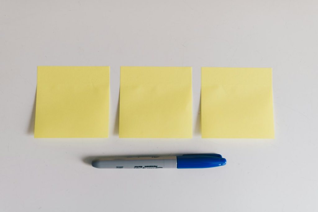 Three blank yellow sticky notes laid in a row on a white desk, with a blue permanent marker resting beneath them, shot from directly above.