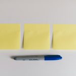 Three blank yellow sticky notes laid in a row on a white desk, with a blue permanent marker resting beneath them, shot from directly above.