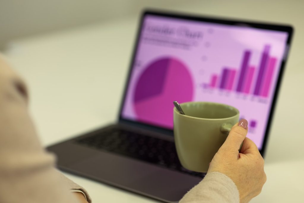 Over-the-shoulder view of a person in a light sweater holding a green ceramic mug in front of an open laptop, its screen showing a magenta and purple analytics dashboard with a pie chart and a rising bar chart, softly out of focus.