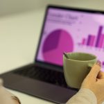 Over-the-shoulder view of a person in a light sweater holding a green ceramic mug in front of an open laptop, its screen showing a magenta and purple analytics dashboard with a pie chart and a rising bar chart, softly out of focus.