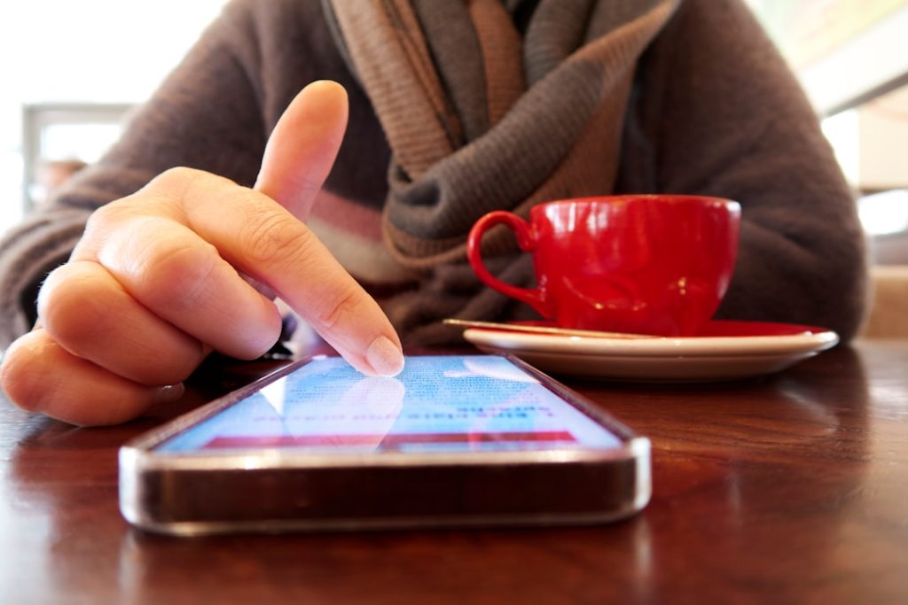 A person's index finger reaching down to tap the screen of a smartphone lying flat on a polished wooden cafe table, a red espresso cup and saucer beside it, the person's brown knit sweater blurred in the background.