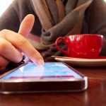 A person's index finger reaching down to tap the screen of a smartphone lying flat on a polished wooden cafe table, a red espresso cup and saucer beside it, the person's brown knit sweater blurred in the background.