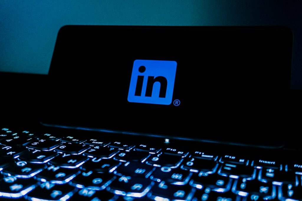 The blue LinkedIn logo glowing on a dark phone screen propped behind a backlit laptop keyboard in a dim room.