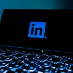 The blue LinkedIn logo glowing on a dark phone screen propped behind a backlit laptop keyboard in a dim room.