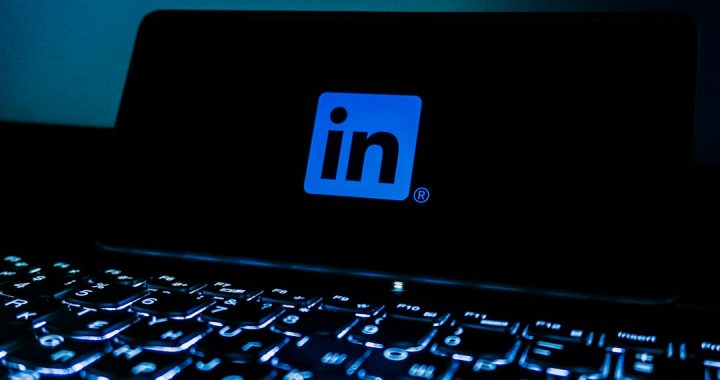 The blue LinkedIn logo glowing on a dark phone screen propped behind a backlit laptop keyboard in a dim room.