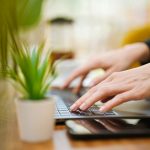 Close-up of a person's hands typing on a laptop keyboard at a wooden table, a small potted green plant in the foreground and a blurred bright room behind