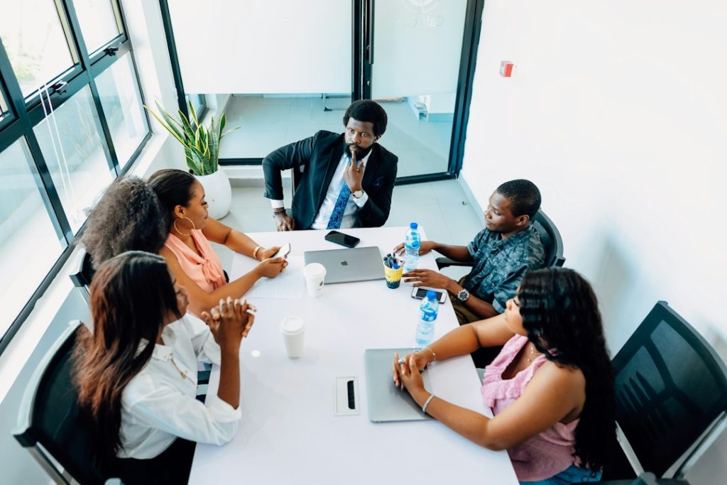 Five colleagues seated around a white conference table in a bright office, one man in a suit resting his hand on his chin while the group listens, laptops, phones and water bottles on the table.