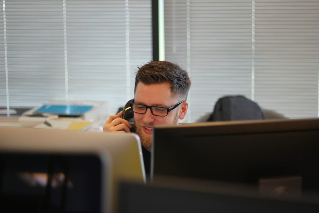A smiling man with glasses and a beard talks on a desk phone in an office, seen between two computer monitors, with window blinds behind him.