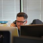 A smiling man with glasses and a beard talks on a desk phone in an office, seen between two computer monitors, with window blinds behind him.