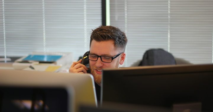 A smiling man with glasses and a beard talks on a desk phone in an office, seen between two computer monitors, with window blinds behind him.