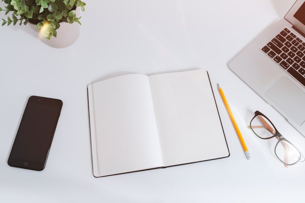 Overhead view of a white desk with an open blank notebook in the center, a yellow pencil and black-rimmed glasses beside it, a dark smartphone on the left, a small green plant in a white pot at top left, and the corner of a silver laptop keyboard at top right.