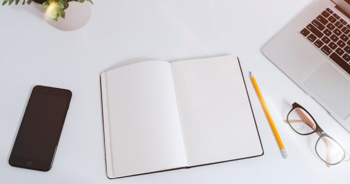 Overhead view of a white desk with an open blank notebook in the center, a yellow pencil and black-rimmed glasses beside it, a dark smartphone on the left, a small green plant in a white pot at top left, and the corner of a silver laptop keyboard at top right.