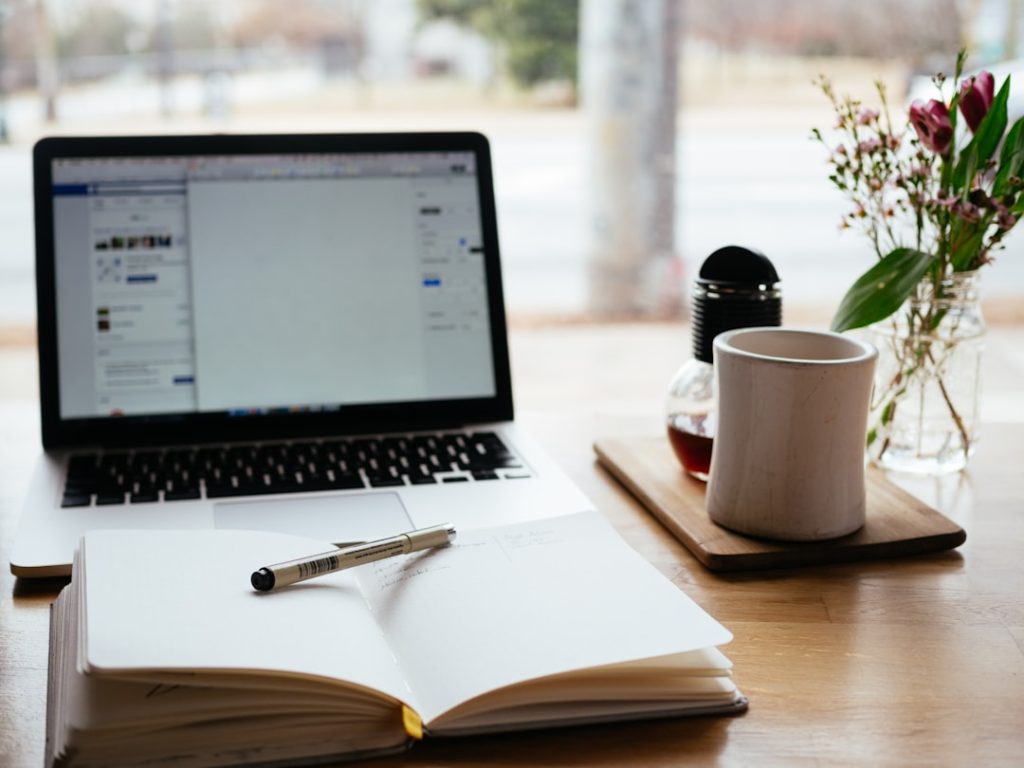 An open notebook with a pen laid across it sits in front of an open laptop on a wooden cafe table, the laptop screen showing a blurred social feed, with a white coffee cup and a jar of pink flowers beside them.