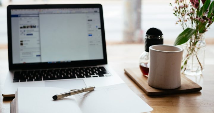 An open notebook with a pen laid across it sits in front of an open laptop on a wooden cafe table, the laptop screen showing a blurred social feed, with a white coffee cup and a jar of pink flowers beside them.