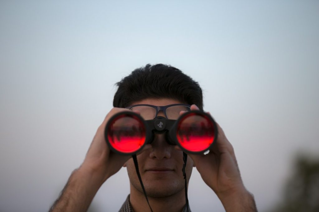 Person with dark hair and glasses holding black binoculars up to their eyes, red-tinted lenses facing the camera, against a pale dusk sky