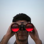 Person with dark hair and glasses holding black binoculars up to their eyes, red-tinted lenses facing the camera, against a pale dusk sky