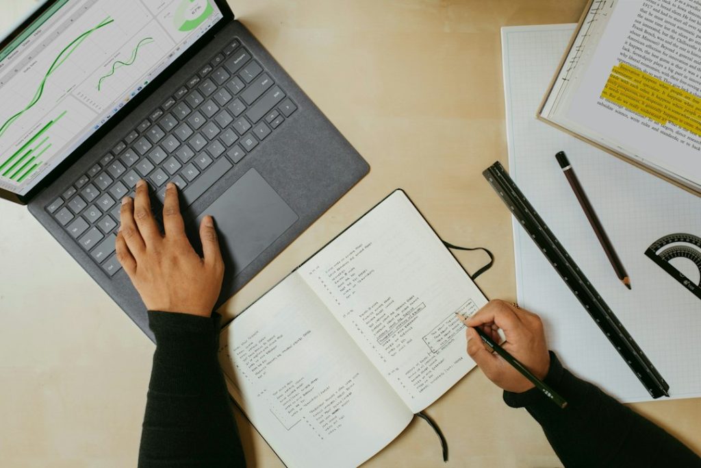 Overhead view of a person's hands at a light wooden desk, one on a dark laptop keyboard showing green line and bar charts, the other writing a list in an open notebook beside a pencil, ruler and an open book with a highlighted paragraph