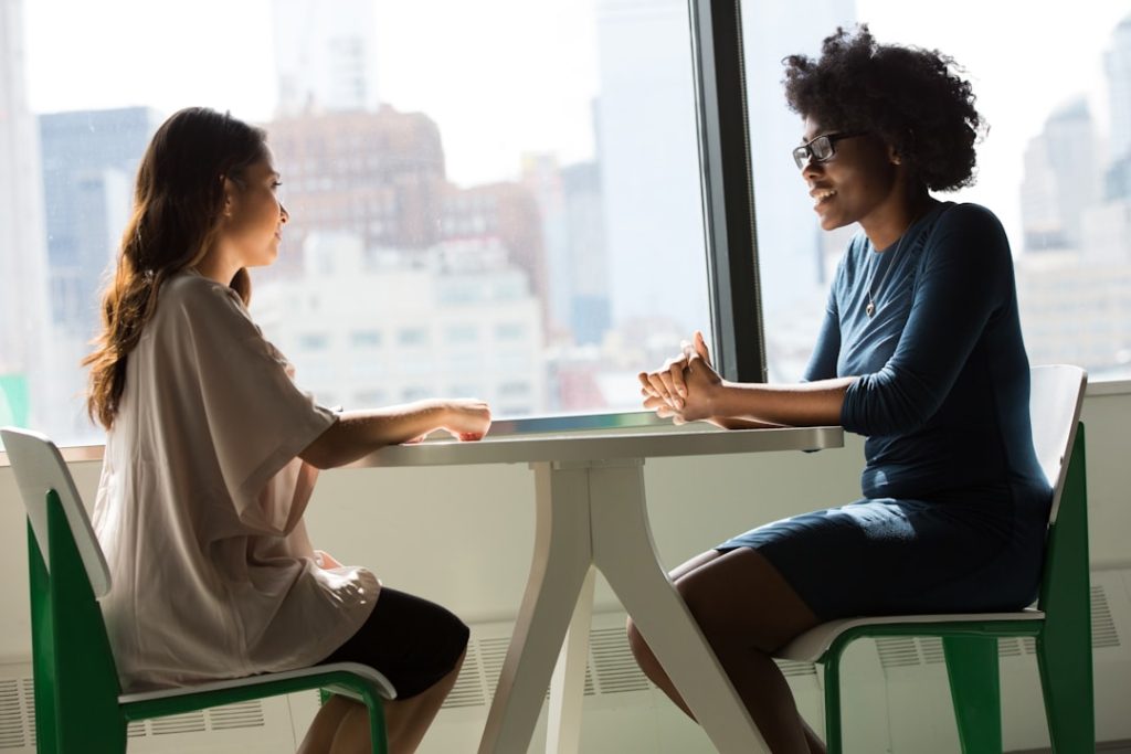 Two people sitting across a small round table beside a floor-to-ceiling office window, talking face to face, with a city skyline visible behind them.